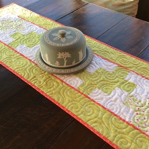 May include: A green and white quilted table runner with a red border. The runner is decorated with a swirl pattern and has a white and green ceramic dish with a lid on top.