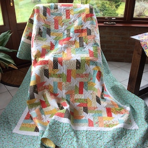 May include: A colourful patchwork quilt with a floral pattern and a teal border. The quilt is folded and draped over a chair in a sunroom.