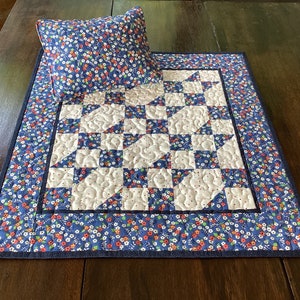 May include: A blue and white quilted table runner with a floral print border. The runner has a white centre with a blue and white geometric pattern. A matching cushion is included.