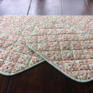May include: A quilted kitchen mat and two matching oven mitts. The set features a light green background with a pattern of various baked goods in shades of pink, brown, and white. The edges are trimmed with a light green binding.