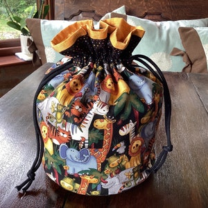 May include: A black drawstring bag with a yellow trim and a colourful print of various animals, including a giraffe, zebra, elephant, and tiger. The bag is sitting on a wooden table.