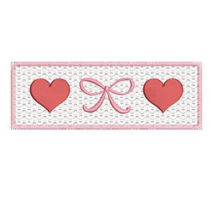 Puede incluir: Un parche bordado con dos corazones y un lazo. Los corazones son rojos y el lazo es rosa. El parche está rodeado por un borde rosa.