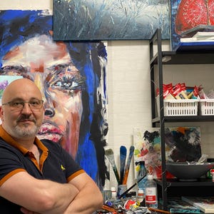 Könnte beinhalten: Ein Mann mit Bart und Brille steht vor einem Gemälde eines Frauenkopfes. Er trägt ein schwarz-oranges Hemd und hat die Arme verschränkt. Im Hintergrund sind weitere Gemälde und Kunstmaterialien zu sehen.