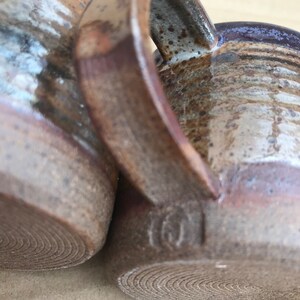 以下が含まれることがあります： 手作りの陶器マグカップ2つ。ざらざらとした土っぽい外観。茶色とベージュのカラーパレットで、濃い紫色の縁とハンドルが付いています。底には渦巻き模様があります。1つのマグカップには小さな刻印があります。