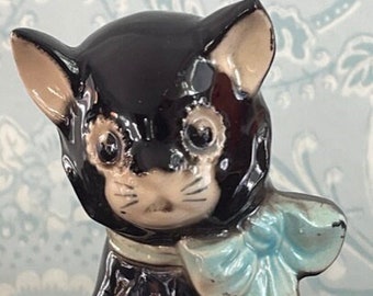 May include: A black ceramic figurine of a cat wearing a blue bow. The cat has a white face and a black nose.