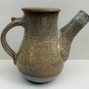 May include: A small, hand-thrown ceramic teapot with a light brown glaze and a white speckled finish. The teapot has a rounded body, a short spout, and a handle that curves up and over the top. The base of the teapot is glazed in a light blue color.