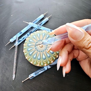 May include: A close-up of a hand using a blue and white dotting tool to create a mandala design on a round, white, green, and blue stone. The stone is surrounded by several other dotting tools.
