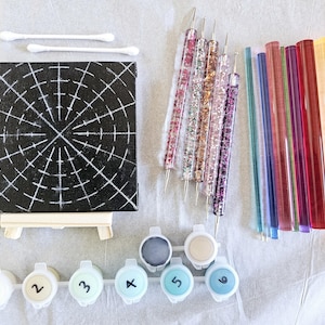 May include: A paint by numbers kit with a black canvas featuring a white grid pattern, six small paint pots labelled 1 through 6, and a set of tools including a set of dotting tools and a set of glass rods.