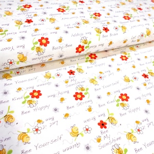 May include: White fabric with a repeating pattern of yellow bees, red flowers, and text. The text includes phrases like "Bee Happy," "Bee Kind," and "Bee Yourself." The fabric is rolled, showing the pattern's continuity.