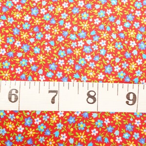 May include: Red fabric with a small floral print in blue, white, and yellow. A white measuring tape with black numbers is placed on top of the fabric. The numbers on the tape read 5, 6, 7, 8, 9.