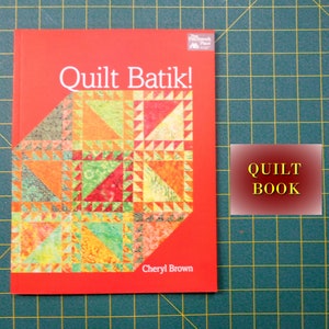 May include: A red book titled "Quilt Batik!" by Cheryl Brown. The cover features a colorful quilt pattern with a variety of triangles in shades of orange, yellow, green, and red. The book is labeled "Quilt Book" in a yellow rectangle.