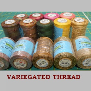 May include: A collection of spools of variegated thread in various colors, including brown, green, pink, and yellow. Each spool is wrapped in clear plastic and has a blue label. The text "VARIEGATED THREAD" is displayed at the bottom.