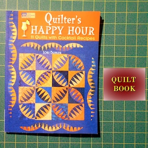 May include: A blue book titled "Quilter's Happy Hour" with a colorful quilt design on the cover. The book features 11 quilts with cocktail recipes and is written by Lori Buhler. The book is labeled "Quilt Book" in a yellow rectangle.