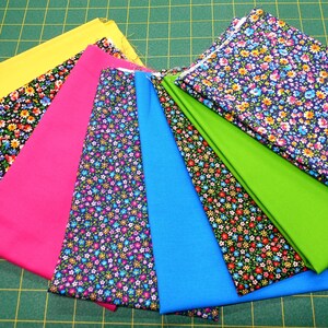 May include: A collection of fabric swatches in various colors and patterns, including yellow, pink, blue, green, and black with white floral designs. The fabrics are arranged in a fan shape on a green cutting mat.