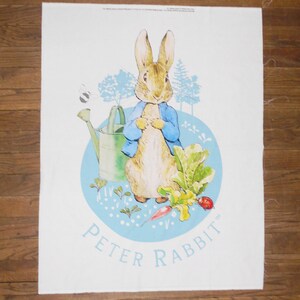 May include: A white fabric panel featuring a printed illustration of Peter Rabbit wearing a blue jacket. The scene includes a green watering can, leafy vegetables, a small bee, and light blue trees set against a circular blue background.