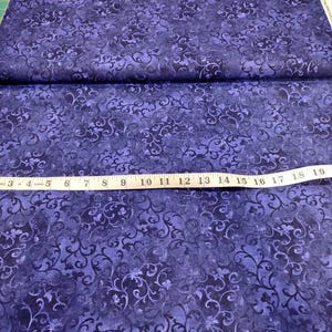 May include: A dark purple fabric featuring a repeating black swirling vine and floral pattern. A white measuring tape is placed horizontally across the fabric, showing a length measurement extending past 22 inches on the scale.