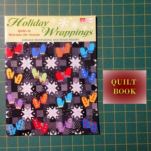May include: A book titled "Holiday Wrappings: Quilts to Welcome the Season" by Loraine Manwaring and Susan Nelsen. The cover features a colorful quilt design with mittens and stars on a black and white background. The book is labeled "Quilt Book".