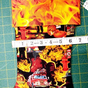 May include: Firefighter themed Christmas stocking with a red fire truck, fire hydrant, and flames. The stocking is made of fabric and has a red, yellow, and black color scheme. The stocking is 6 inches long.