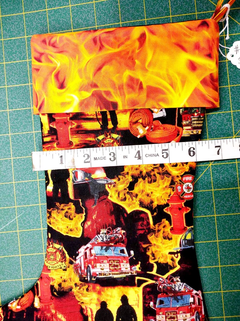 May include: Firefighter themed Christmas stocking with a red fire truck, fire hydrant, and flames. The stocking is made of fabric and has a red, yellow, and black color scheme. The stocking is 6 inches long.