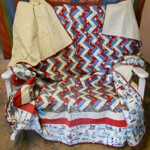 May include: A red, white, and blue chevron patterned quilt with a nautical theme. The quilt is folded and draped over a white rocking chair. The quilt features a border with lighthouses and seashells.