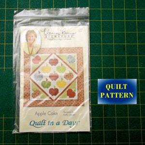 May include: A quilt pattern titled "Apple Cake" from the "Quilt in a Day" series by Eleanor Burns Signature. The pattern features a quilt design with red, green, and yellow apples on a white background.