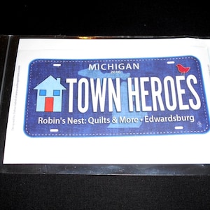 May include: A blue and white Michigan license plate with the text "Town Heroes" and a red bird. The plate also includes the text "Robin's Nest: Quilts & More - Edwardsburg".