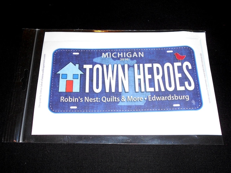 May include: A blue and white Michigan license plate with the text "Town Heroes" and a red bird. The plate also includes the text "Robin's Nest: Quilts & More - Edwardsburg".