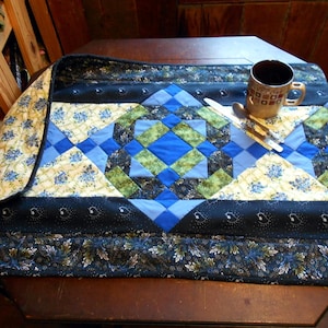 May include: A blue and green patchwork table runner with a black border and a floral pattern. A brown coffee mug with a white design sits on the runner.