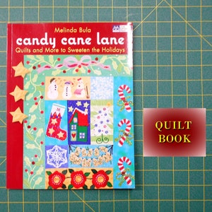 May include: A red book titled "candy cane lane" by Melinda Bula. The book cover features a patchwork design with a variety of Christmas-themed illustrations, including candy canes, snowmen, stockings, and a house. The book's subtitle is "Quilts and More to Sweeten the Holidays".