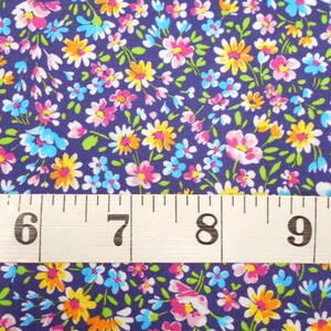May include: A close-up of a purple fabric with a small floral print in shades of pink, yellow, blue, and white. A white measuring tape with black numbers is placed on top of the fabric, showing the numbers 6, 7, 8, and 9.