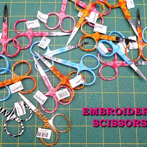 May include: A collection of colorful embroidery scissors with decorative patterns. The scissors have a variety of designs, including floral, geometric, and animal prints. The text "Embroidery Scissors" is printed in pink letters on a green background.
