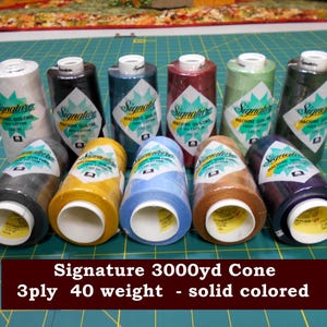 May include: A collection of ten spools of Signature brand machine quilting thread in various solid colors. The spools are labeled with the brand name and product details. The text at the bottom reads: Signature 3000yd Cone, 3ply 40 weight - solid colored.
