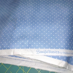 May include: Light blue fabric with a white polka dot pattern. The fabric has a white border with the text "Beautiful Basics by Maywood Studio © Copyright". The fabric is folded and appears to be new.