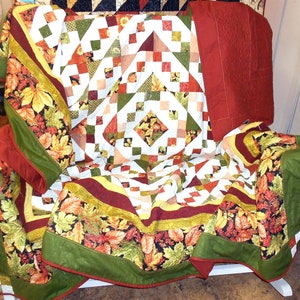May include: A patchwork quilt with a white background and a variety of colorful squares and diamonds. The quilt features a fall leaf pattern in shades of green, brown, and orange. The quilt is draped over a chair.
