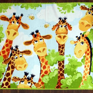 May include: A colorful fabric panel featuring a whimsical design of five giraffes with brown and orange spots, surrounded by green leaves and blue sky. The giraffes have large, expressive eyes and friendly smiles.