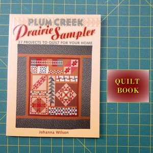 May include: A book titled "Plum Creek Prairie Sampler" with a colorful quilt design on the cover. The book has a yellow background and the title is in brown and red lettering. The subtitle is "27 Projects to Quilt for Your Home" and the author is Johanna Wilson. The book is labeled "Quilt Book".