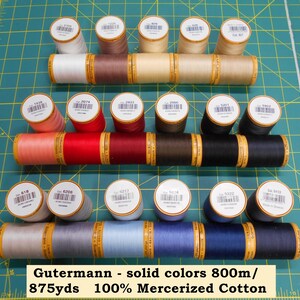 May include: A collection of Gutermann solid color thread spools in various shades, including white, brown, red, black, and blue. The spools are arranged on a green cutting mat. The text "Gutermann - solid colors 800m/875yds 100% Mercerized Cotton" is visible.