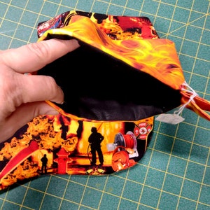 May include: A small black fabric pouch with a zipper closure. The pouch is lined with black fabric and has a fire-themed print on the outside. The print features flames, firefighters, and a fire hydrant. The pouch is open and a hand is holding the top of the pouch.