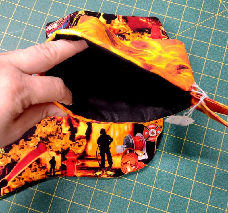 May include: A small black fabric pouch with a zipper closure. The pouch is lined with black fabric and has a fire-themed print on the outside. The print features flames, firefighters, and a fire hydrant. The pouch is open and a hand is holding the top of the pouch.