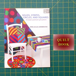 May include: A book titled "Solids, Stripes, Circles, and Squares" with 16 modern patchwork quilt patterns by Pippa Eccles Armbrester. The cover features a colorful illustration of a chair, table, and rug with geometric patterns.