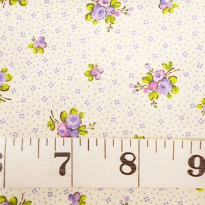 May include: White fabric with a repeating pattern of small purple flowers and a larger cluster of purple flowers with green leaves. A white measuring tape with black numbers 6, 7, 8, and 9 is placed at the bottom of the image.
