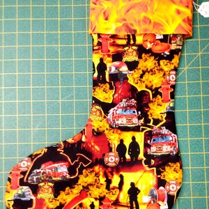 May include: A red and yellow Christmas stocking with a fire department theme. The stocking features images of firefighters, fire trucks, fire hydrants, and flames. The stocking has a red cuff with a yellow flame pattern.