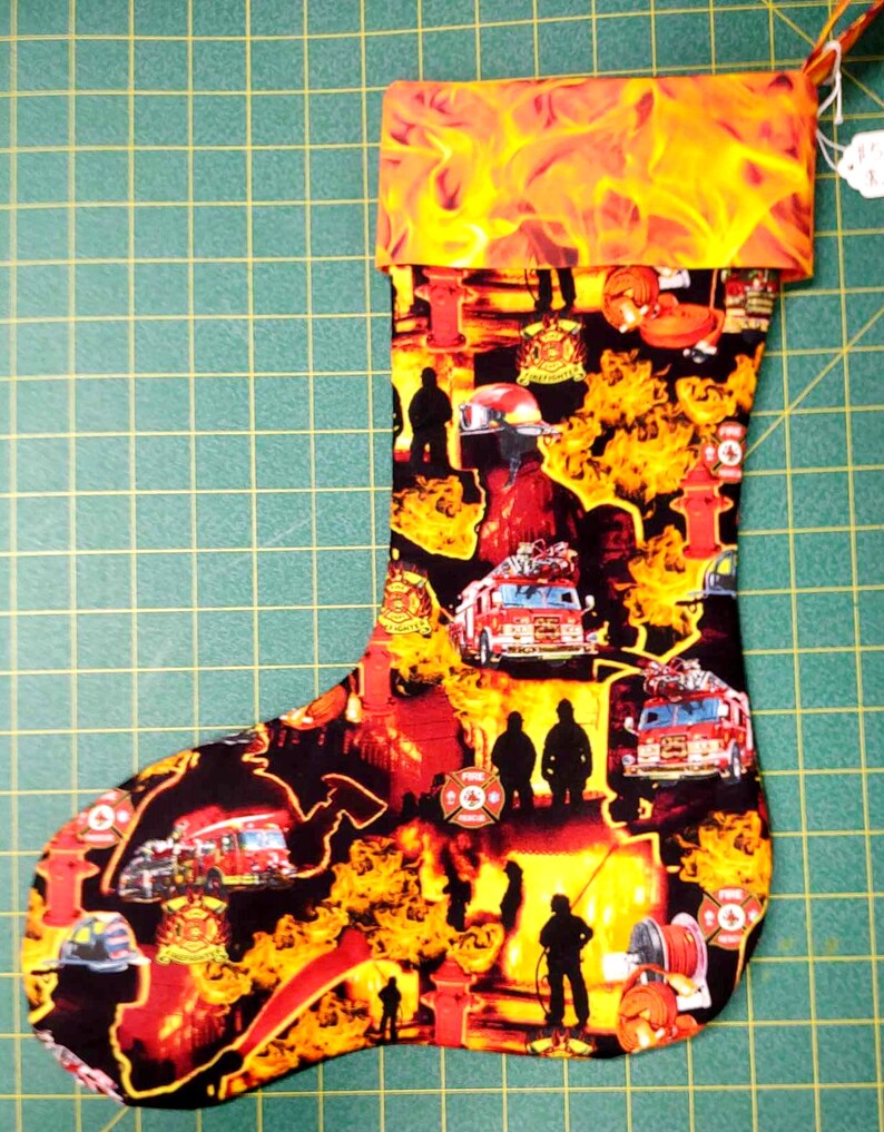 May include: A red and yellow Christmas stocking with a fire department theme. The stocking features images of firefighters, fire trucks, fire hydrants, and flames. The stocking has a red cuff with a yellow flame pattern.