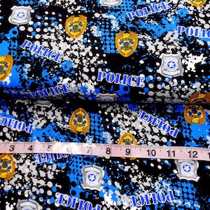 May include: Blue and black fabric with a repeating pattern of police badges, white paint splatters, and the word "POLICE" in blue. The fabric is measured with a tape measure.