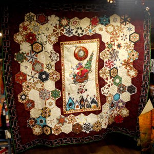 May include: A handmade Christmas quilt with a central panel depicting Santa Claus in a sleigh with a Christmas tree. The quilt features a burgundy border and a pattern of hexagonal shapes with snowflake and star designs.