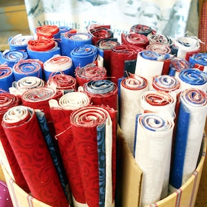 May include: A container filled with rolled fabric in red, white, and blue. The fabric rolls are various sizes and patterns, including swirls and patriotic designs. The container has a striped pattern.