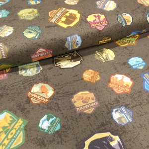May include: Dark gray fabric featuring colorful patches with national park names and illustrations. Patches include Yellowstone, Rocky Mountain, and Zion National Park. The fabric has a textured, mottled appearance.