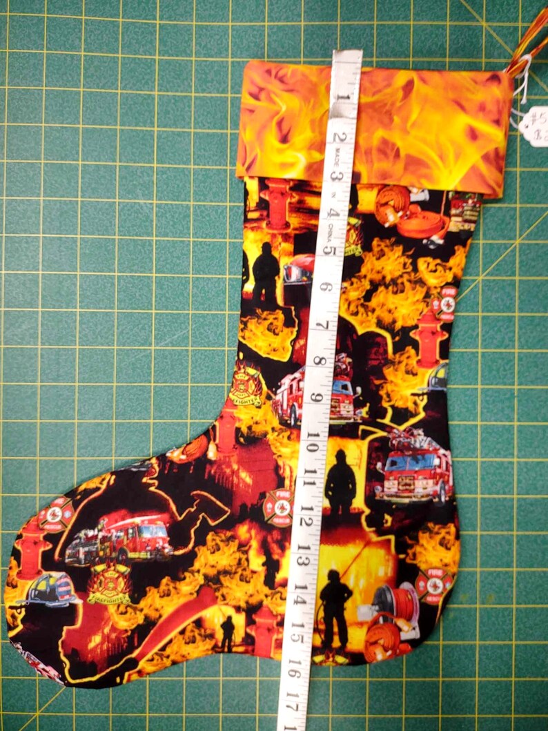 May include: A red and yellow Christmas stocking with a fire truck and firefighter theme. The stocking is 17 inches long and has a cuff with flames.