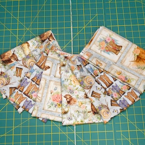 May include: Several fabric squares with a light blue background and a Western theme. The fabric features images of cowboy boots, hats, flowers, and a longhorn. The fabric is arranged on a green cutting mat.