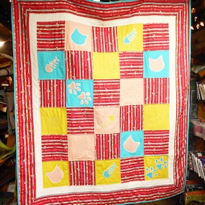 May include: A handmade quilt with a patchwork design featuring cat-themed motifs. The quilt has a red and white striped border and a white inner border. The squares are in shades of red, yellow, pink, and teal, with cat faces, fish bones, and paw prints.